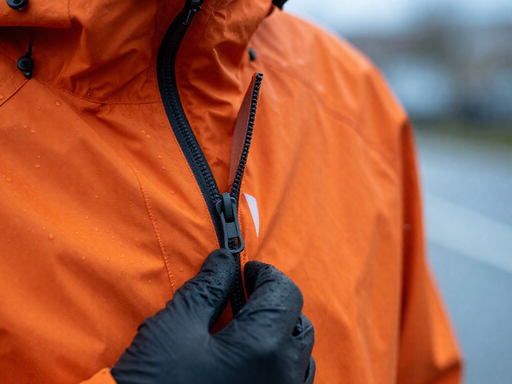 Close-up of a hand zipping a bright orange rain jacket, showing sealed seams and a waterproof zipper with water droplets on the fabric.