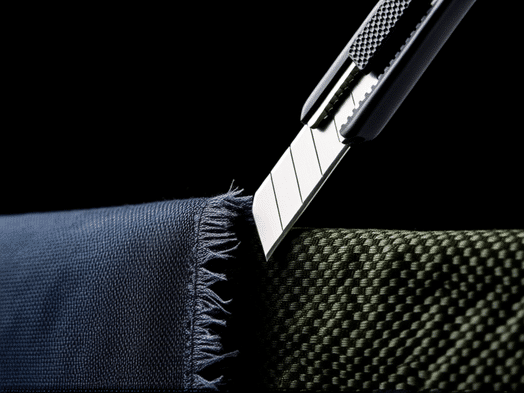 Extreme detail, a box cutter blade pressed against two different fabrics. One is generic nylon, fraying instantly. The other