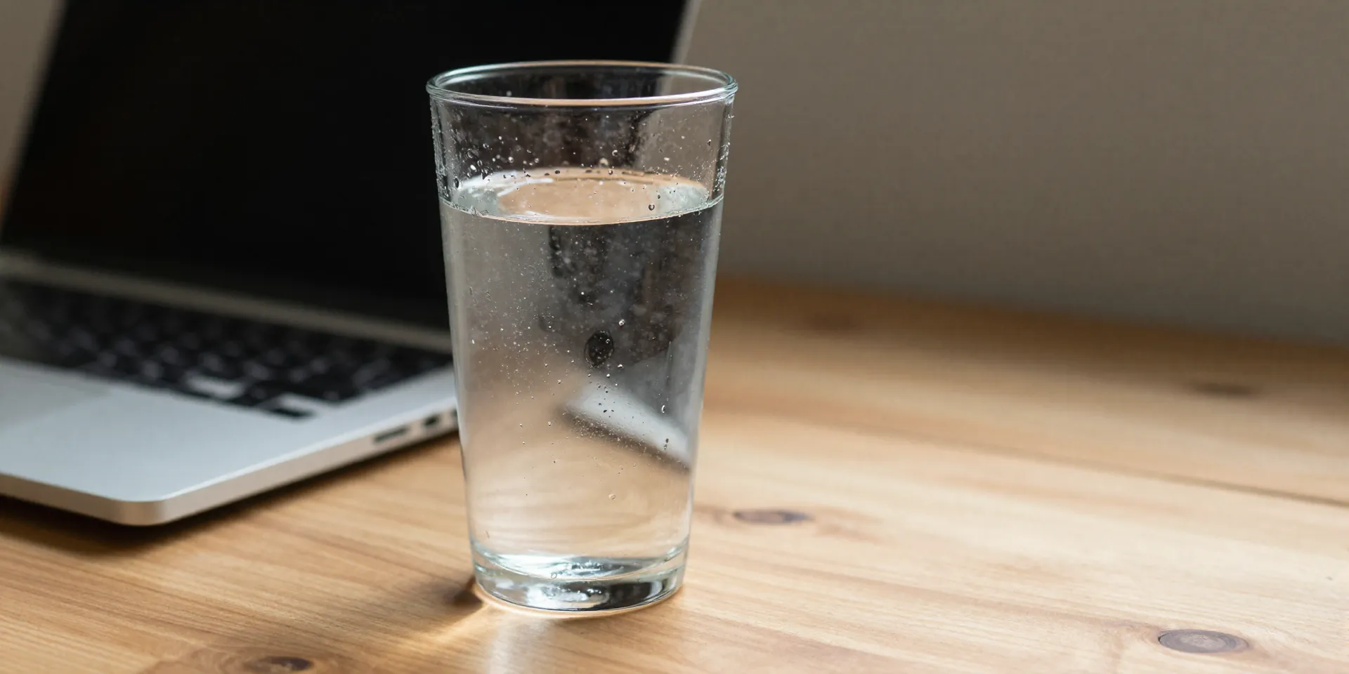 midjourney prompt: close-up photo of a clear, tall glass of water on a wooden desk next to a laptop, morning light, condensation on the glass, visually refreshing, cinematic shallow focus --style raw --ar 4:5