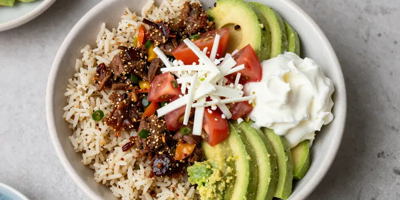 Overhead shot of a beautifully composed keto Chipotle bowl: cilantro-lime cauliflower rice topped with carnitas, fajita veggies, fresh tomato salsa, cheese, guacamole, and sour cream. Vibrant colors, rustic texture, natural light food photo.