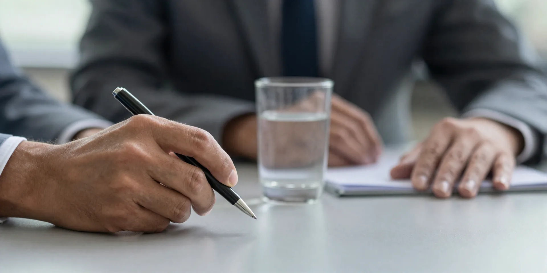 Close-up of a calm, focused executive's hands on a conference table during a tense meeting. One hand is loosely gripping a sleek black fountain pen, the other rests calmly near a glass of water. Ambient office light, sense of quiet control. --ar 4:3