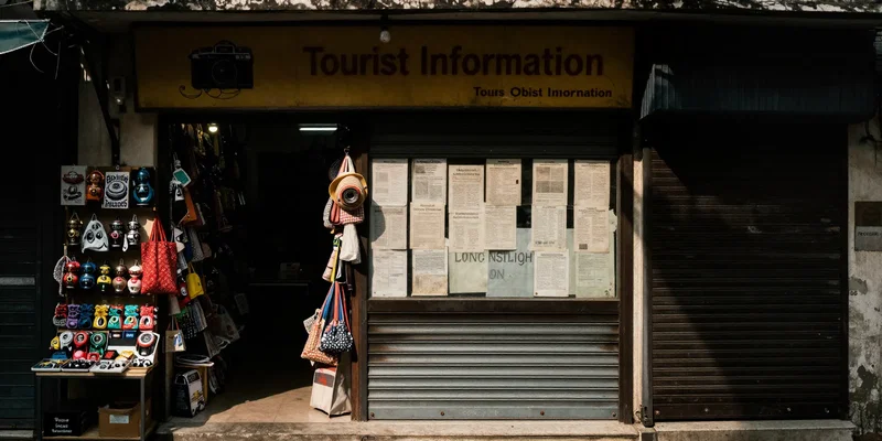 A dim, narrow storefront in Hanoi's Old Quarter labeled 'Tourist Information,' crammed between a souvenir shop and a closed shutter, with faded printed A4 papers taped to the window. Cinematic lighting, deep shadows, shot on a vintage lens.