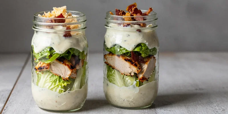 mason jar filled with layers of parmesan, grilled chicken, romaine, and a creamy dressing, bacon crumbles and parmesan crisp visible at the top, side light, rustic kitchen setting, sharp focus