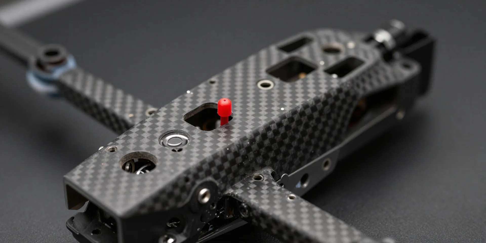 Macro photography of a carbon fiber FPV drone frame with a small red antenna awkwardly touching the frame plates, shallow depth of field, sharp focus, workshop lighting --ar 16:9