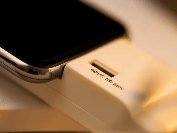 A close-up of a modern smartphone and laptop charger, with tiny text 'INPUT: 100-240V' clearly visible and highlighted. Macro lens, shallow depth of field, warm light.