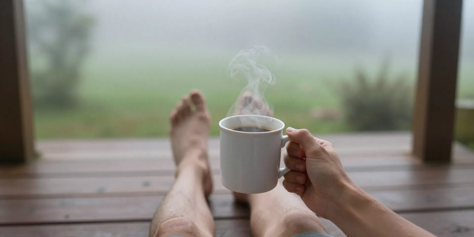First-person perspective from a porch or garden. Bare feet on wooden decking, a slight morning fog, a steaming mug of coffee