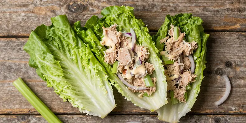 Top-down flat lay, style of a food blog, two crisp iceberg lettuce leaves used as wraps, filled with chunky keto tuna salad (tuna, mayo, celery, red onion). Rustic wood background, fresh ingredients scattered around.