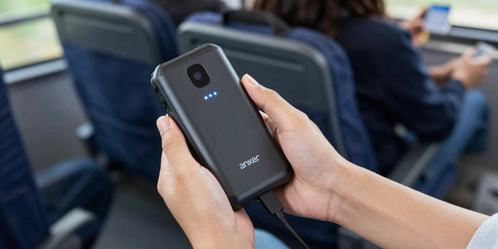 Action shot. A person's hands holding a specific, popular rugged power bank (like an Anker PowerCore Elite) with a glowing LED power indicator. The background is the blurry, moving interior of a bus, with other passengers and bags out of focus. The cable is plugged into a phone. Sense of motion and utility. --ar 16:9