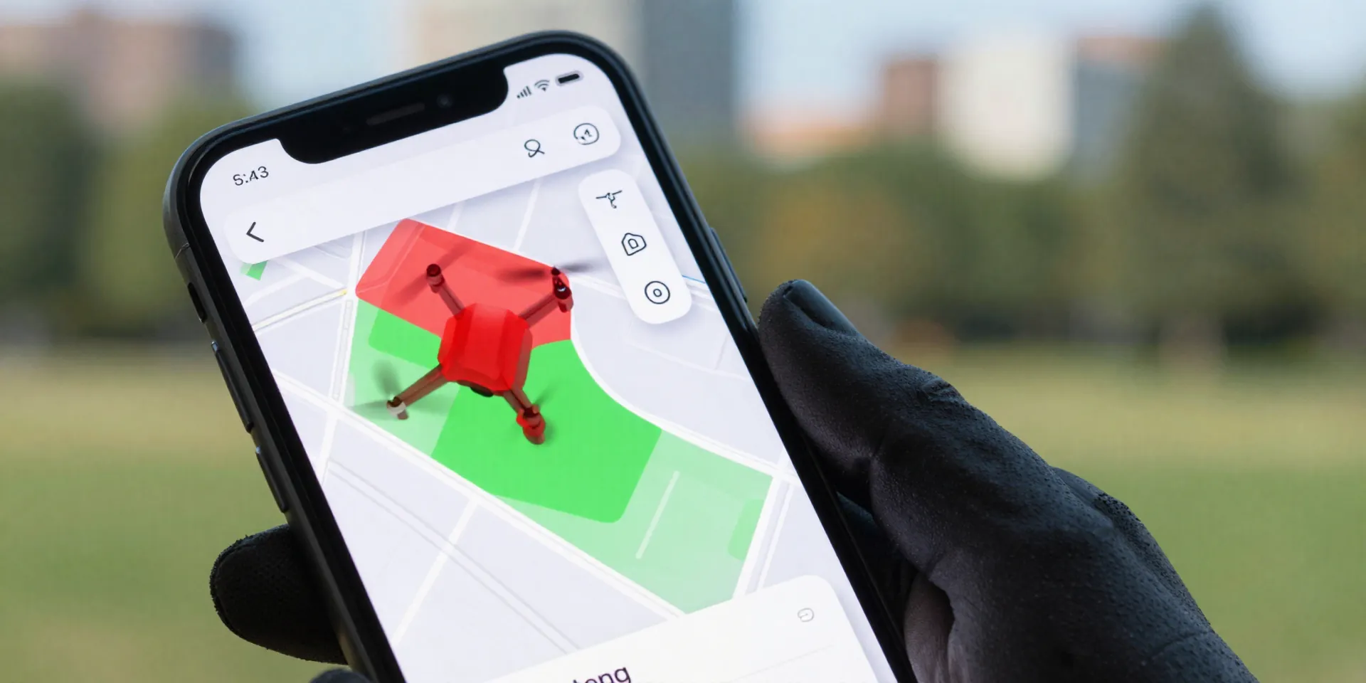 Close up of a smartphone screen showing a drone map app with red and green flight zones, held by a hand wearing fingerless gloves, blurred urban park background, photorealistic, depth of field --ar 16:9